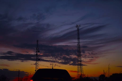 Two communication towers rise against a backdrop of a dramatic sunset sky filled with vibrant and dark clouds. A silhouetted vehicle is visible in the foreground, and the sky transitions from deep blues to fiery oranges and reds near the horizon.