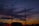 Two communication towers rise against a backdrop of a dramatic sunset sky filled with vibrant and dark clouds. A silhouetted vehicle is visible in the foreground, and the sky transitions from deep blues to fiery oranges and reds near the horizon.