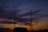 Two communication towers rise against a backdrop of a dramatic sunset sky filled with vibrant and dark clouds. A silhouetted vehicle is visible in the foreground, and the sky transitions from deep blues to fiery oranges and reds near the horizon.