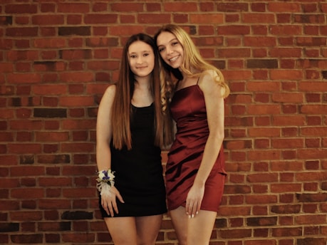 Two young women are standing side by side against a red brick wall. One is wearing a black dress with long hair and a floral wrist corsage. The other is wearing a dark red dress with wavy blonde hair.