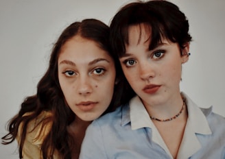 Two young women are closely positioned, both looking directly at the camera. The woman on the left has long, wavy hair, and wears a light-colored top, while the woman on the right has short, dark hair and wears a collared shirt with a beaded necklace. The background is plain and neutral.
