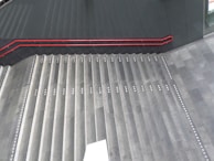 A set of wide, concrete stairs with a red handrail on the left side. The steps are equipped with white tactile paving dots, likely for aiding visually impaired individuals. The floor and the surrounding walls are made of dark materials. A white rectangular object appears at the bottom center of the stairs.