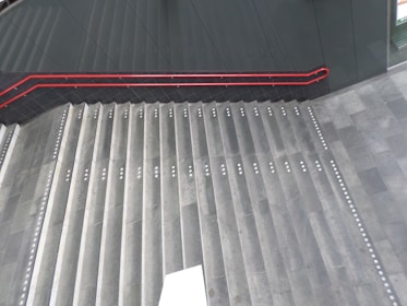 A set of wide, concrete stairs with a red handrail on the left side. The steps are equipped with white tactile paving dots, likely for aiding visually impaired individuals. The floor and the surrounding walls are made of dark materials. A white rectangular object appears at the bottom center of the stairs.