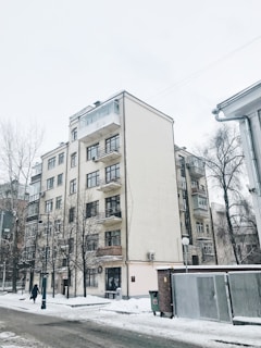 A multi-story residential building with balconies is seen, set against a snowy urban landscape. Bare trees and street lamps line the street, with a person walking along the snowy pathway. The sky is overcast, adding to the winter atmosphere.