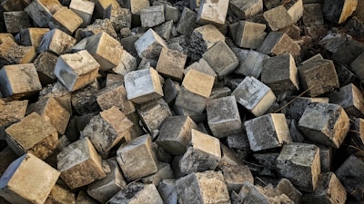 A large pile of rough, textured concrete blocks arranged haphazardly, creating a chaotic and rugged appearance. The blocks are predominantly cubic in shape, with uneven, worn surfaces reflecting various shades of gray.