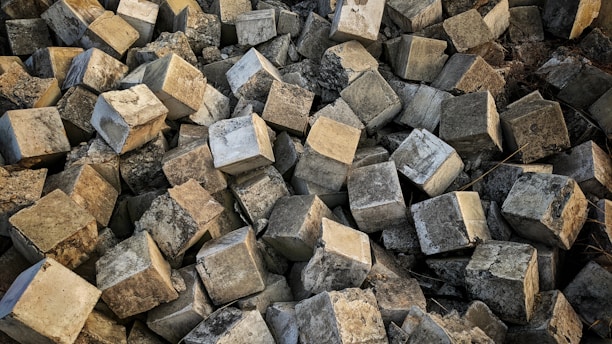 A large pile of rough, textured concrete blocks arranged haphazardly, creating a chaotic and rugged appearance. The blocks are predominantly cubic in shape, with uneven, worn surfaces reflecting various shades of gray.