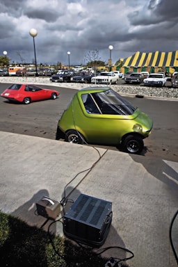 A small, compact green car is parked on a paved area, connected by cables to a nearby device. In the background, a red car is visible on the road, and several parked cars can be seen behind a fence. The sky is overcast with dark clouds, and a yellow-and-green striped tent is set up in the distance.