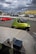 A small, compact green car is parked on a paved area, connected by cables to a nearby device. In the background, a red car is visible on the road, and several parked cars can be seen behind a fence. The sky is overcast with dark clouds, and a yellow-and-green striped tent is set up in the distance.
