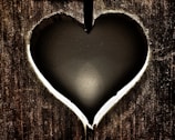 A heart-shaped cutout in a wooden surface, revealing a dark, smooth background behind it. The edges of the heart cutout are illuminated with light, creating a glowing outline.