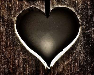 A heart-shaped cutout in a wooden surface, revealing a dark, smooth background behind it. The edges of the heart cutout are illuminated with light, creating a glowing outline.
