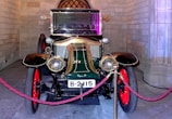 A vintage car with a polished brass finish on its headlights and grille, featuring red spoked wheels and a glossy green and black body. It is displayed indoors against a backdrop of stone walls, and surrounded by a decorative red rope barrier.