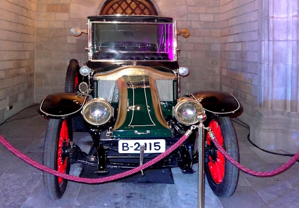 A vintage car with a polished brass finish on its headlights and grille, featuring red spoked wheels and a glossy green and black body. It is displayed indoors against a backdrop of stone walls, and surrounded by a decorative red rope barrier.