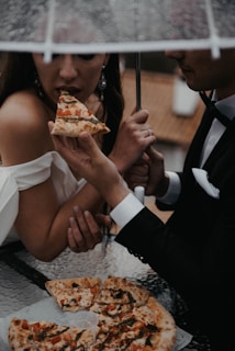 A couple dressed elegantly in formal attire is sharing a slice of pizza under a transparent umbrella. The woman is taking a bite while holding the umbrella, and the man's hand is seen holding the pizza slice. The setting seems intimate and romantic, with a table and more pizza slices visible.