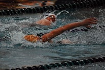 Two swimmers are in a competitive swimming pool, performing the freestyle stroke. They are partially submerged, surrounded by splashing water, with their arms outstretched. The pool has lane dividers visible.