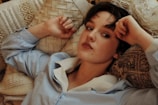 A person with short dark hair is reclining on an assortment of textured pillows and cushions. They wear a light blue shirt with a white collar and have a relaxed yet contemplative expression.