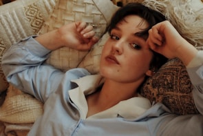 A person with short dark hair is reclining on an assortment of textured pillows and cushions. They wear a light blue shirt with a white collar and have a relaxed yet contemplative expression.
