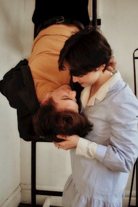 A couple is sharing an intimate moment. One person is upside down, possibly hanging from a structure, while the other supports and kisses them tenderly. The setting appears to be indoors, with minimalistic decor.