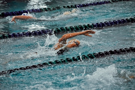 Several swimmers in action, performing the front crawl stroke in a swimming pool with lane dividers visible. Water splashes around them as they swim intensely.
