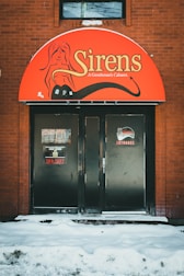 A brick building facade with a red awning featuring the name 'Sirens: A Gentleman's Cabaret' along with a graphic illustration of a woman. The entrance consists of double black doors, one with an 'Open' sign partially visible and the word 'Entrance' written in red. Snow covers the ground in front of the entrance.