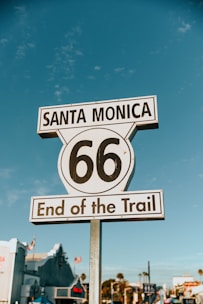A road sign with the text 'Santa Monica 66 End of the Trail' against a clear blue sky. In the background, there are indistinct buildings and palm trees.