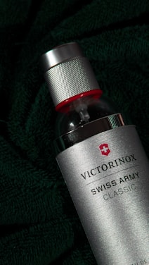 A silver spray bottle with a textured metal cap and red accent on top. The bottle is labeled 'Victorinox Swiss Army Classic' and is set against a dark green, textured fabric background, adding a sense of luxury and sophistication.