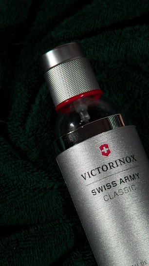 A silver spray bottle with a textured metal cap and red accent on top. The bottle is labeled 'Victorinox Swiss Army Classic' and is set against a dark green, textured fabric background, adding a sense of luxury and sophistication.