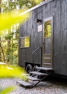 A modern, black tiny house with a single door and a small staircase leading up to it is located in a wooded area. The house features a sleek design with a few windows and is mounted on wheels. The surrounding environment is lush with greenery, and there is a picnic table nearby.