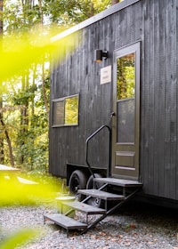 A modern, black tiny house with a single door and a small staircase leading up to it is located in a wooded area. The house features a sleek design with a few windows and is mounted on wheels. The surrounding environment is lush with greenery, and there is a picnic table nearby.