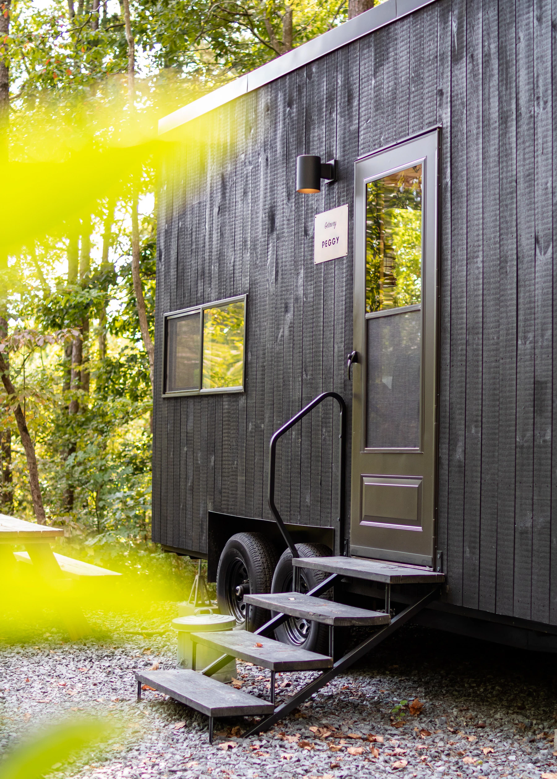 The door, fold-down metal steps and twin trailer wheels of a black-sided tiny house standing on gravel