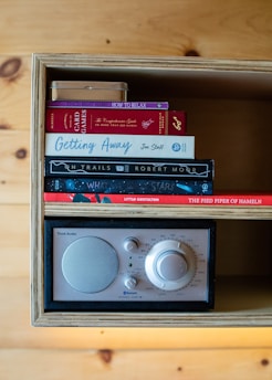 A wooden shelf houses an assortment of books stacked on top of each other alongside a retro-style radio. The book titles include 'How to Relax', 'The Comprehensive Guide to More Than 350 Games', and 'Getting Away'. The radio features knobs and a speaker grille, suggesting an analog design with a touch of modernity through Bluetooth functionality. The warm tones of the wood complement the colorful book spines.