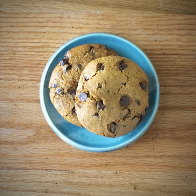 Two chocolate chip cookies are placed on a light blue ceramic plate, which rests on a wooden surface. The cookies have a golden brown appearance and are generously studded with chocolate chips.