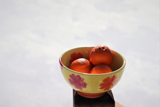 A floral-patterned bowl containing three bright orange tangerines sits in natural daylight. The bowl features red and pink flowers painted on its yellow surface and is placed on a wooden object with a light, slightly blurred background.