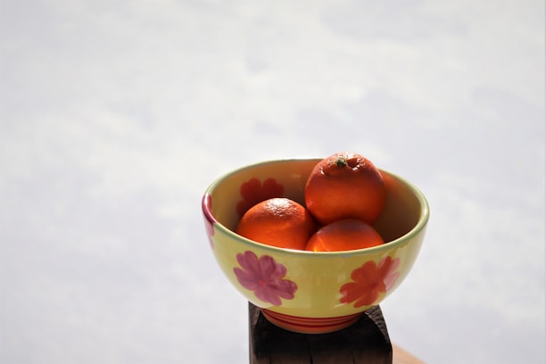 A floral-patterned bowl containing three bright orange tangerines sits in natural daylight. The bowl features red and pink flowers painted on its yellow surface and is placed on a wooden object with a light, slightly blurred background.