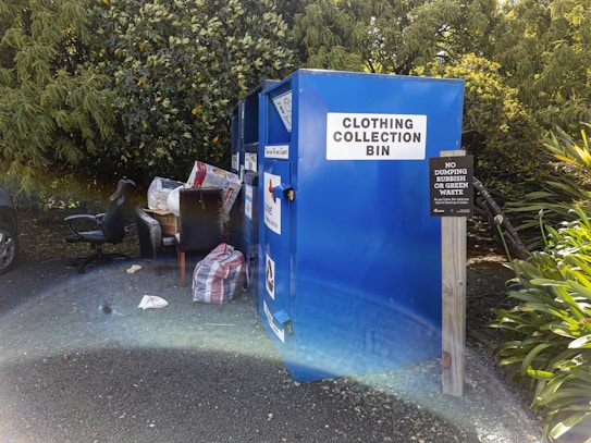 A blue clothing collection bin stands outdoors near a cluster of lush green trees. On the ground next to the bin, there are various discarded items including a black office chair, a striped bag, and a large box. A sign adjacent to the bin warns against dumping rubbish or green waste.
