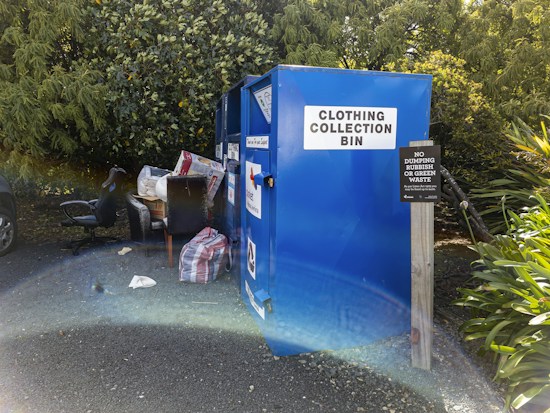 A blue clothing collection bin stands outdoors near a cluster of lush green trees. On the ground next to the bin, there are various discarded items including a black office chair, a striped bag, and a large box. A sign adjacent to the bin warns against dumping rubbish or green waste.
