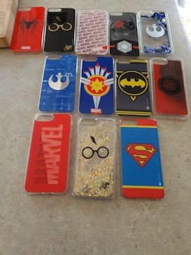 A collection of twelve phone cases displayed on a flat surface. The designs feature various superhero and pop culture symbols, including Spider-Man, Harry Potter glasses and lightning bolt, Marvel logos, and symbols associated with Star Wars and other franchises. The cases are predominantly made of plastic with colorful prints and patterns.
