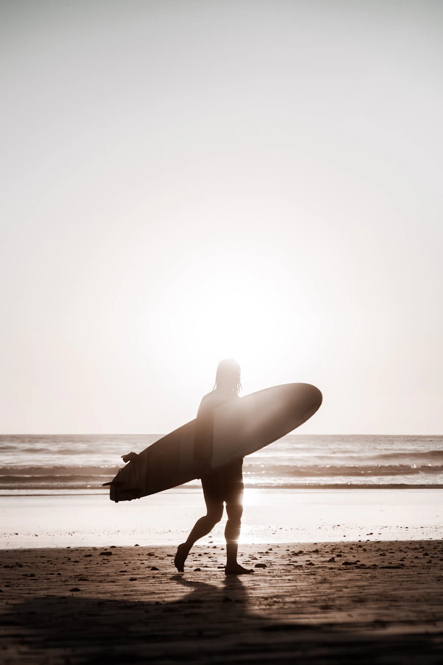 Surfista caminando por la playa cargando una tabla hacia el agua al atardecer