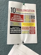 A poster pinned to a textured blue board, titled '10 Decolonisation Actions for Non-Māori Kiwis'. The poster lists various actions such as learning about Māori history, understanding the Declaration of Independence & Te Tiriti o Waitangi, speaking up for treaty settlement processes, strengthening relationships with Māori organizations, and challenging inequities. Each action is highlighted in different blocks of colors including red, yellow, and black, contributing to the visual emphasis of the text.