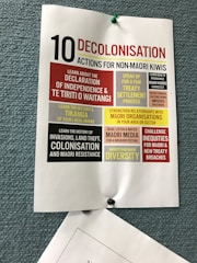 A poster pinned to a textured blue board, titled '10 Decolonisation Actions for Non-Māori Kiwis'. The poster lists various actions such as learning about Māori history, understanding the Declaration of Independence & Te Tiriti o Waitangi, speaking up for treaty settlement processes, strengthening relationships with Māori organizations, and challenging inequities. Each action is highlighted in different blocks of colors including red, yellow, and black, contributing to the visual emphasis of the text.