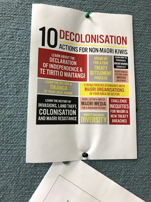 A poster pinned to a textured blue board, titled '10 Decolonisation Actions for Non-Māori Kiwis'. The poster lists various actions such as learning about Māori history, understanding the Declaration of Independence & Te Tiriti o Waitangi, speaking up for treaty settlement processes, strengthening relationships with Māori organizations, and challenging inequities. Each action is highlighted in different blocks of colors including red, yellow, and black, contributing to the visual emphasis of the text.