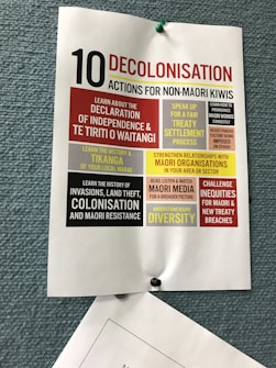 A poster pinned to a textured blue board, titled '10 Decolonisation Actions for Non-Māori Kiwis'. The poster lists various actions such as learning about Māori history, understanding the Declaration of Independence & Te Tiriti o Waitangi, speaking up for treaty settlement processes, strengthening relationships with Māori organizations, and challenging inequities. Each action is highlighted in different blocks of colors including red, yellow, and black, contributing to the visual emphasis of the text.