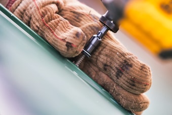 A close-up of a gloved hand holding a power drill next to a green surface, with a screw being driven in. The glove is brown with worn-out spots, and the drill has a metallic black component, with part of it being yellow.
