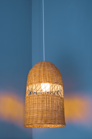 A woven rattan pendant light fixture is suspended from the ceiling against a muted teal wall. The light inside creates a warm, soft glow, highlighting the intricate pattern of the weave.