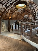 An intricately designed interior space featuring a ceiling woven from natural materials such as tree branches or wooden vines. The floor and railing are crafted in a similar fashion, creating a cohesive rustic aesthetic. Soft, natural lighting filters through gaps in the ceiling, highlighting the organic shapes and textures. Plants adorn an opening in the ceiling, blending indoors with outdoors.