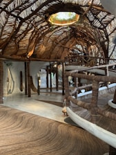 An intricately designed interior space featuring a ceiling woven from natural materials such as tree branches or wooden vines. The floor and railing are crafted in a similar fashion, creating a cohesive rustic aesthetic. Soft, natural lighting filters through gaps in the ceiling, highlighting the organic shapes and textures. Plants adorn an opening in the ceiling, blending indoors with outdoors.