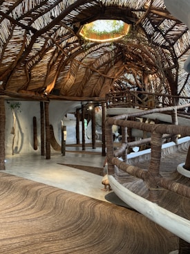 An intricately designed interior space featuring a ceiling woven from natural materials such as tree branches or wooden vines. The floor and railing are crafted in a similar fashion, creating a cohesive rustic aesthetic. Soft, natural lighting filters through gaps in the ceiling, highlighting the organic shapes and textures. Plants adorn an opening in the ceiling, blending indoors with outdoors.