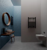A modern bathroom with light blue brick walls, featuring a round wooden mirror, a minimalist wooden shelf with a white vessel sink, and a small potted plant. A towel is placed on a stool under the shelf, and there's a black wall-mounted towel radiator beside a wall-mounted toilet.