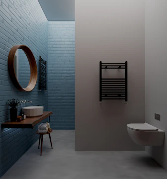 A modern bathroom with light blue brick walls, featuring a round wooden mirror, a minimalist wooden shelf with a white vessel sink, and a small potted plant. A towel is placed on a stool under the shelf, and there's a black wall-mounted towel radiator beside a wall-mounted toilet.