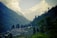 A serene hillside village surrounded by lush green vegetation and tall trees, with rows of quaint houses nestled in the landscape. In the background, majestic mountains peek through a veil of clouds, creating a stunning natural backdrop.