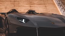 A sleek, modern sports car with a matte black finish is positioned prominently, showcasing its angular design and sharp headlights. The car is set against a backdrop of stone steps, enhancing its luxurious and high-performance appearance.